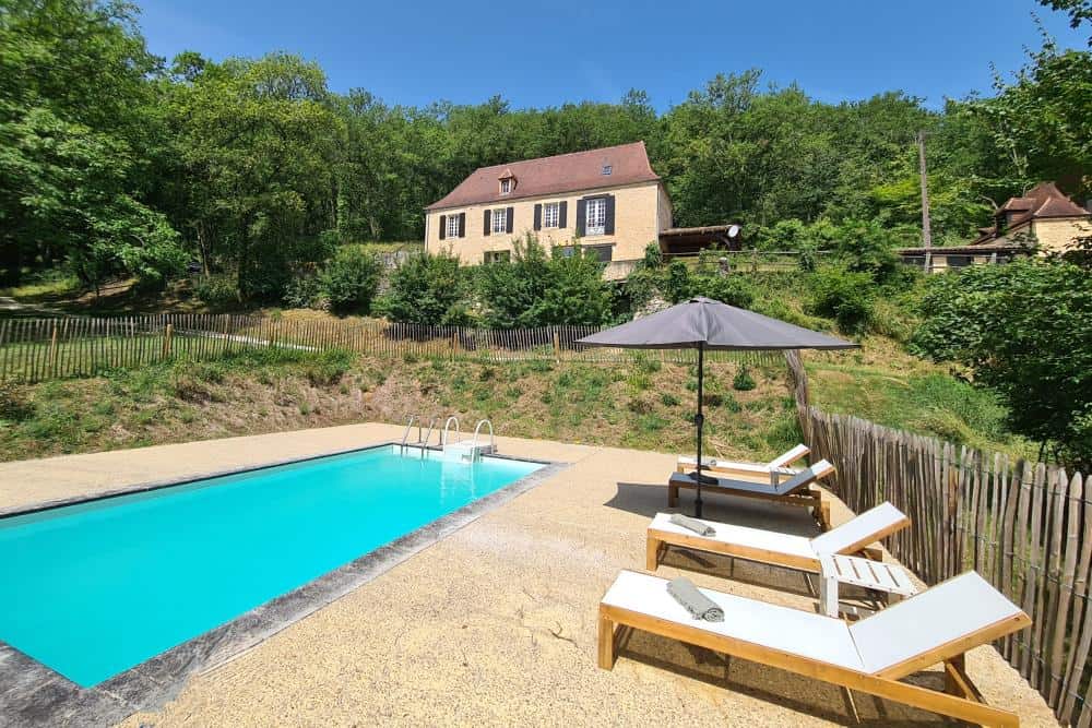 Alt-Text: Schwimmbad im Freien mit Liegestühlen und Sonnenschirm, umgeben von Natur und im Hintergrund ein gemütliches Haus.