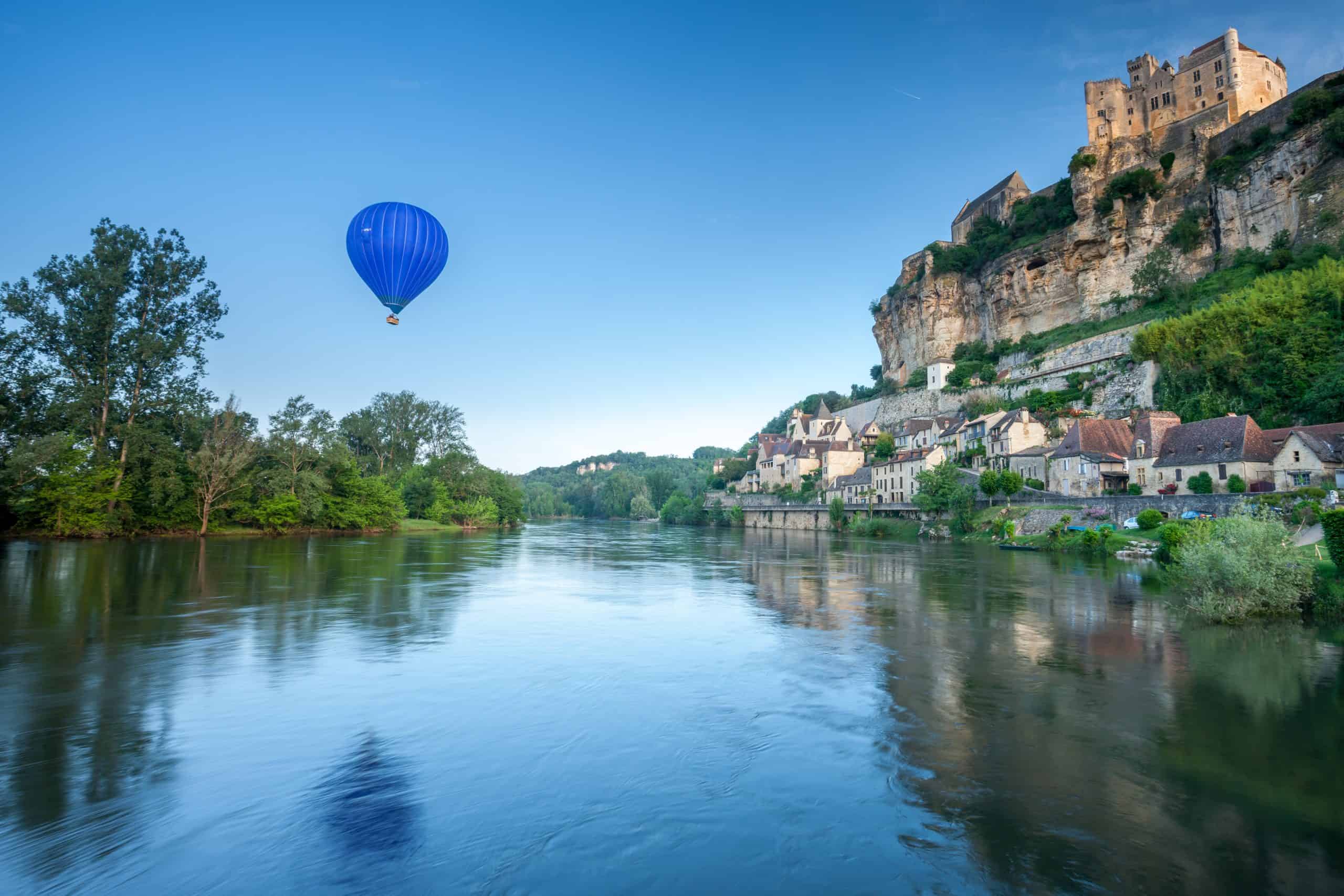 ALT: Charmantes französisches Dorf mit Schloss auf Felsen, Hot-Air-Ballon über Fluss und grüner Umgebung.